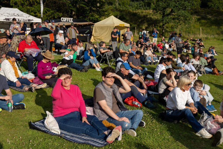 Crowd seated on grassy slope, many covering eyes from sun, at Kenmore Highland Games