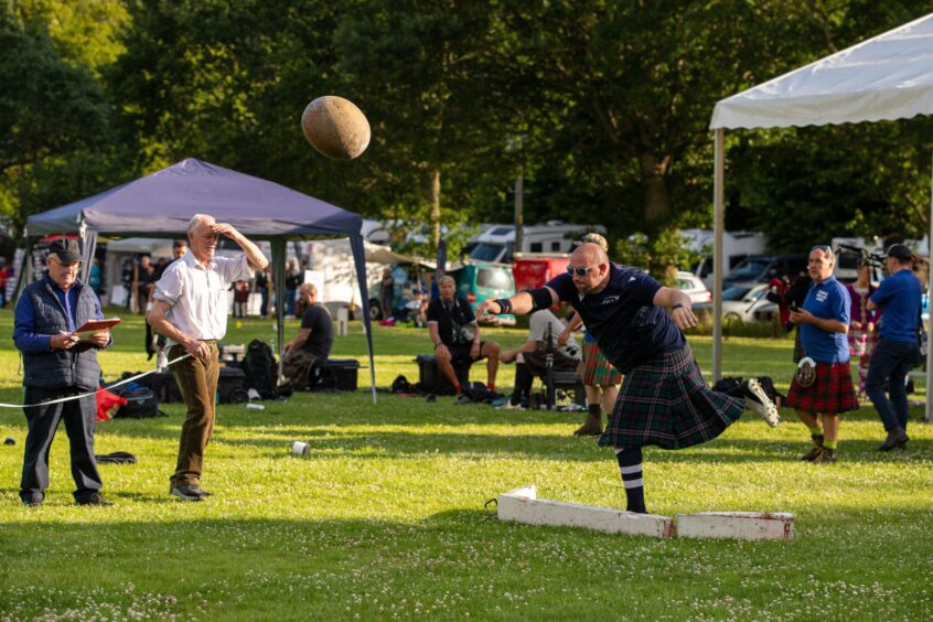Heavy athlete in kilt throwing large round stone watched by judges