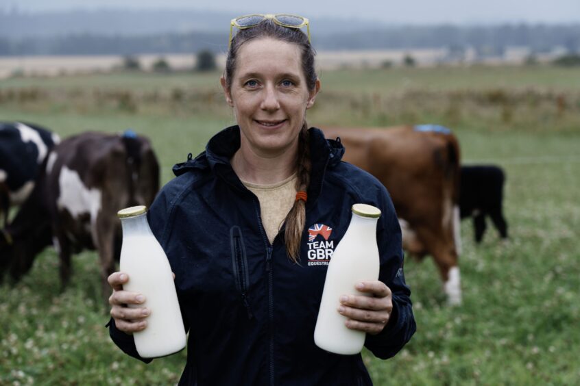 Rachael Cratchley smiling holding two bottles of milk with cows in field behind her