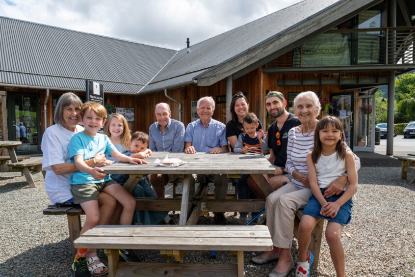 Group of children and adults seated around wooden table outside shop
