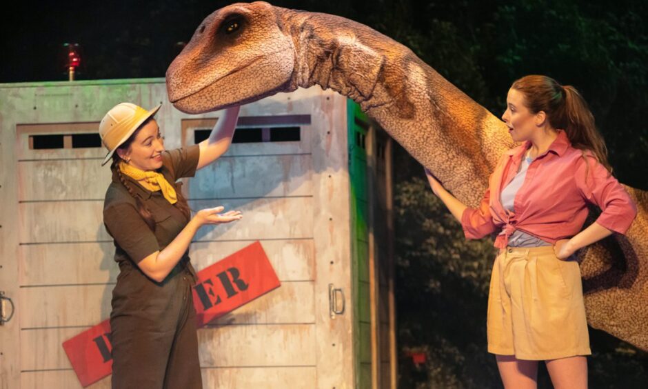 Image shows two actresses dressed in safari/adventure gear with a realistic-looking dinosaur on stage. What's On in Fife.