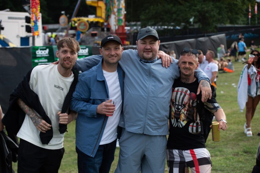 Four men pose together at doof in the Park in Dundee with funfair rides and festival-goers in the background.