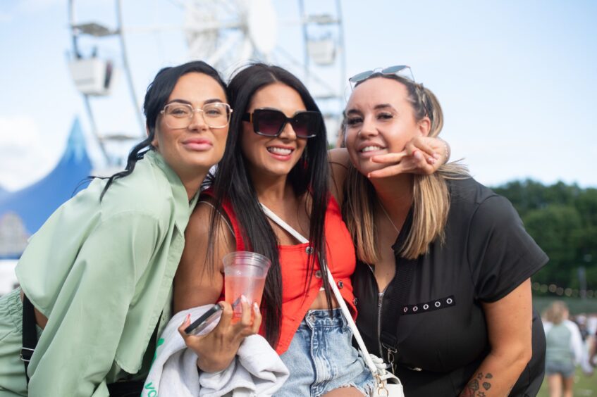Three women smiling and posing with peace signs in front of a Ferris wheel at doof in the Park 2025, Dundee.