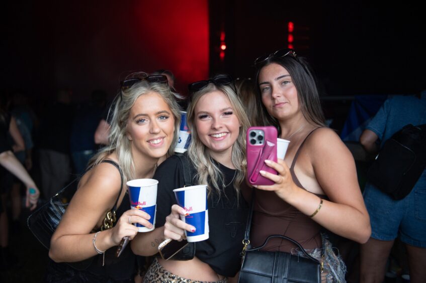 Three women posing for a selfie while holding drinks at doof in the Park 2025, Dundee, with a Red Bull-branded cup in hand.