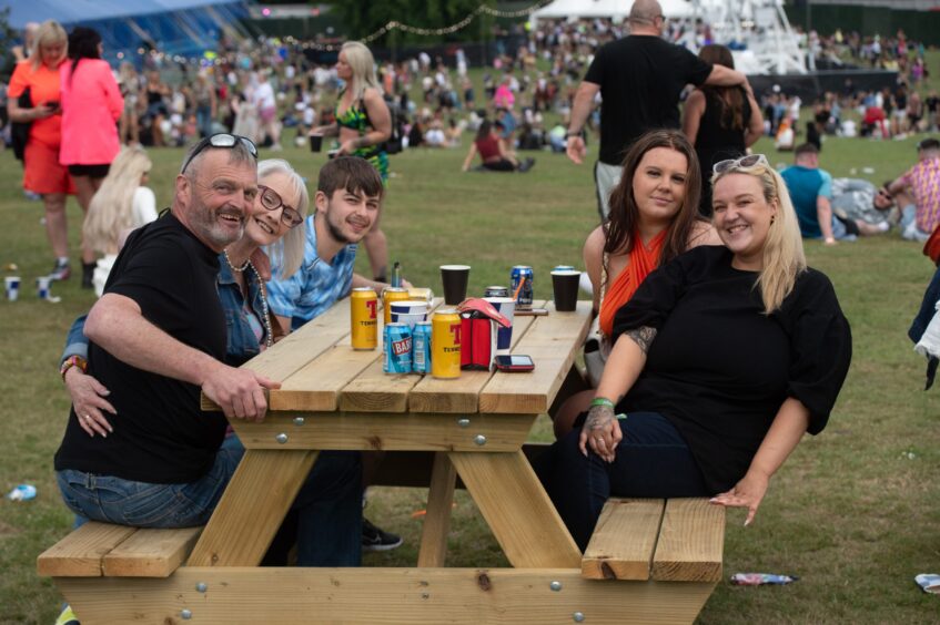 Six people sitting around a wooden picnic bench with drinks, smiling at doof in the Park 2025, Camperdown Park, Dundee.