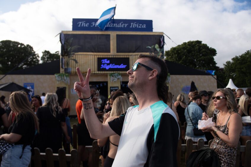 Man in sunglasses poses in front of The Highlander Ibiza as a crowd gathers at doof in the Park 2025 in Dundee.
