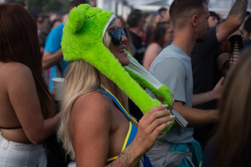 Woman wearing a bright green novelty hat and sunglasses while dancing in the crowd at doof in the Park 2025, Camperdown Park, Dundee.