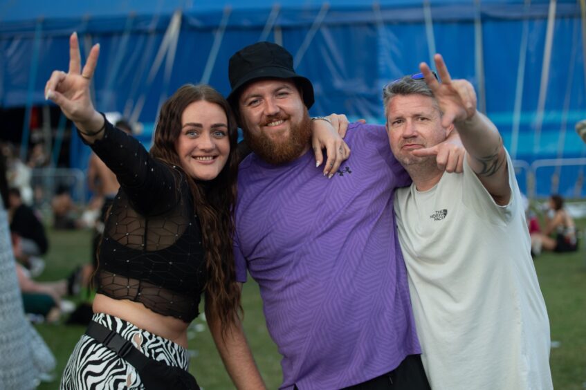 Three friends smiling and flashing peace signs in front of a large blue tent at Doof in the Park 2025, Camperdown Park, Dundee.
