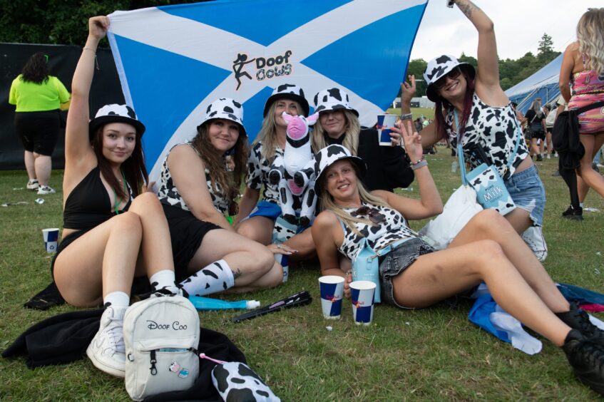 Group of women in cow print outfits with a Scottish flag reading “Doof Cows” at Doof in the Park 2025, Camperdown Park, Dundee.