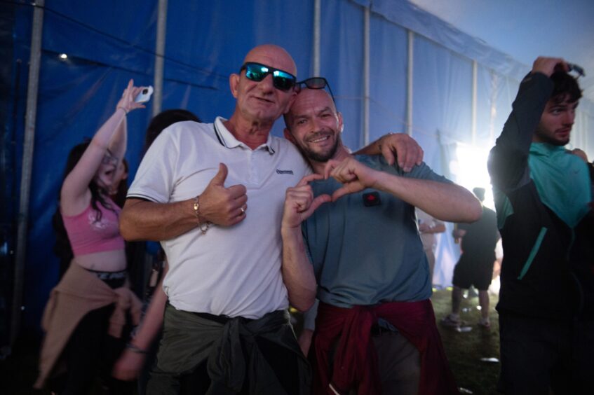 Two men smiling, one giving a thumbs up and the other making a heart shape with hands inside a tent at doof in the Park 2025, Dundee.