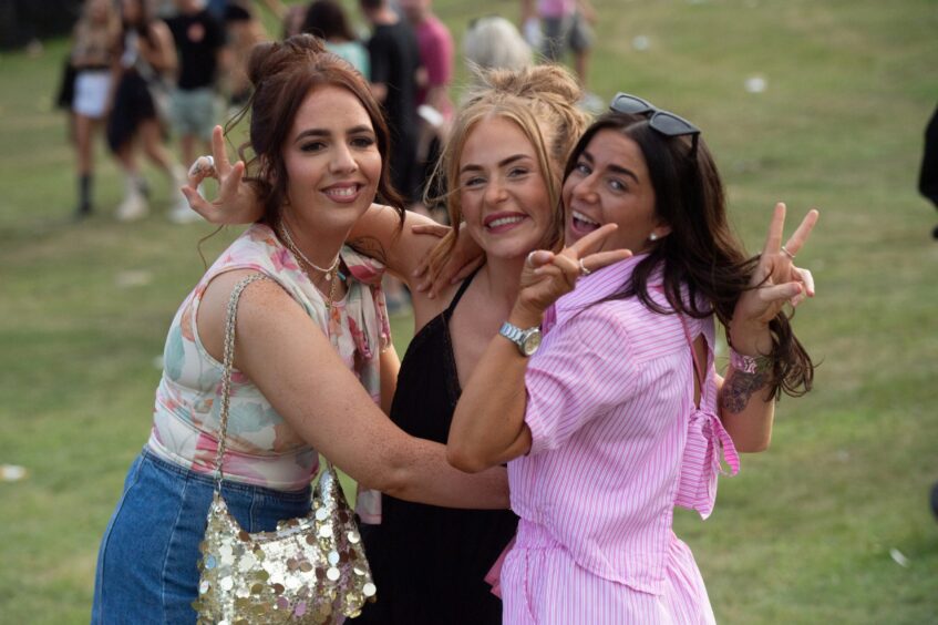 Three women smile and pose with peace signs during doof in the Park 2025 at Camperdown Park, Dundee.