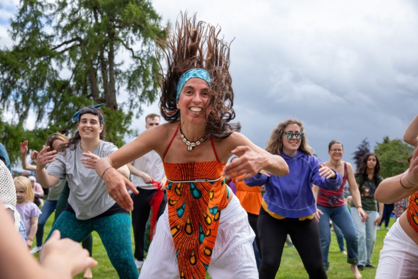 Smiling woman in vibrant orange patterned dress leads a dance session with a crowd at the Afro-Caribbean Summer Festival in Pittencrieff Park, Dunfermline.