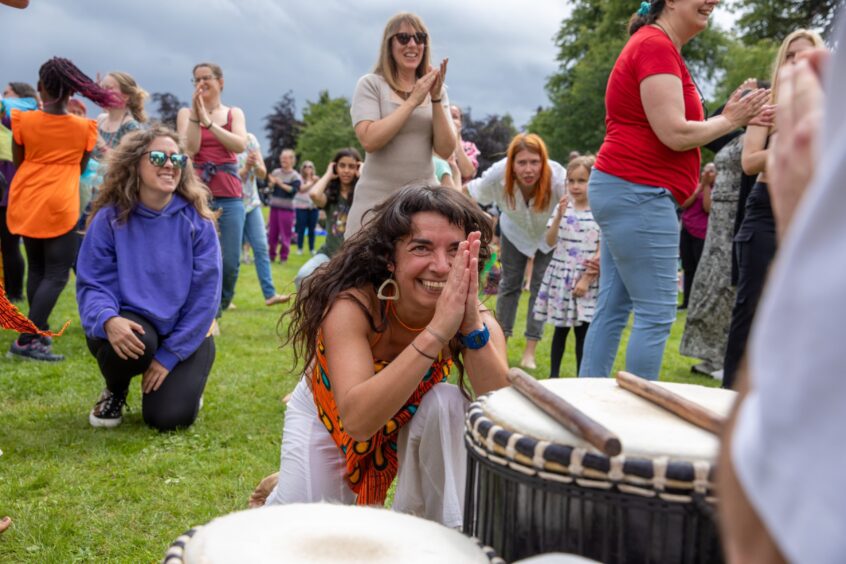Smiling woman in patterned orange outfit kneels beside djembe drums, surrounded by clapping festivalgoers at the Afro-Caribbean Summer Festival in Pittencrieff Park.