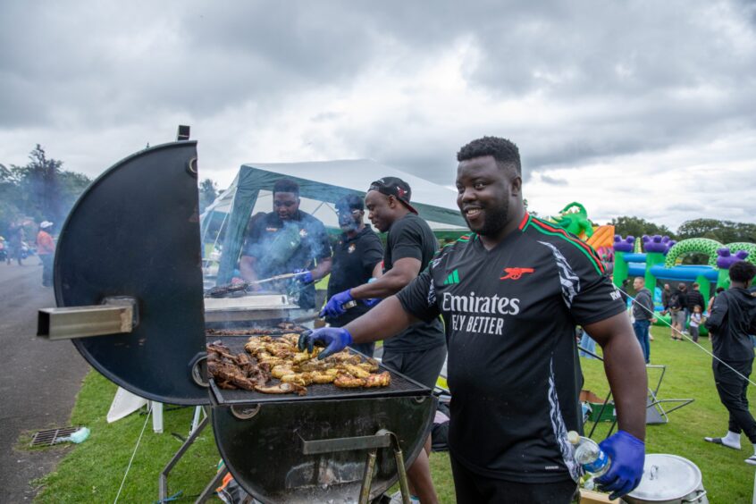 Man grills seasoned meat over a smoky barbecue while smiling, with others cooking beside him at the Afro-Caribbean Summer Festival in Pittencrieff Park.