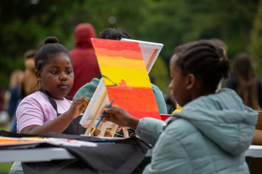 Two children painting vibrant red and yellow sunset scenes on canvases at an arts and crafts table during the Afro-Caribbean Summer Festival in Pittencrieff Park.