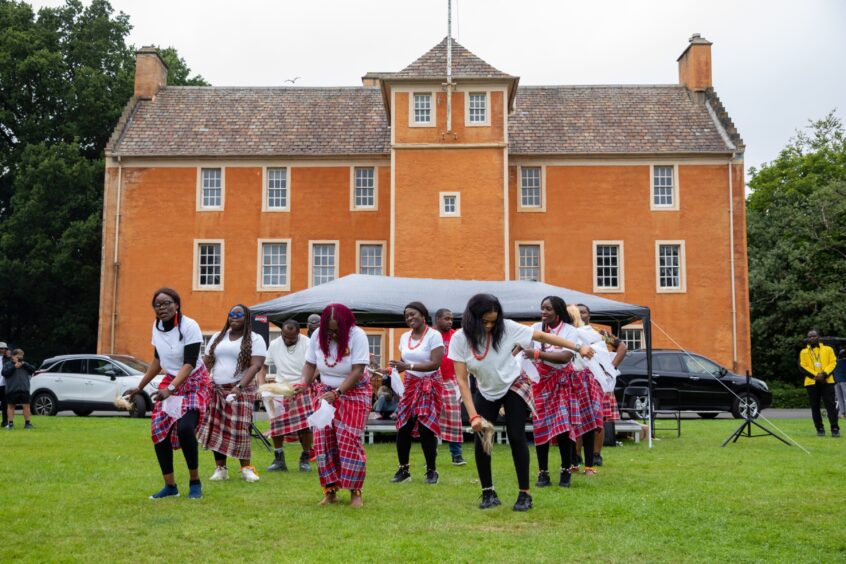 Igbo Amaka Ladies Cultural Group in white tops and red patterned wraps perform a cultural dance outside a large orange building at the Afro-Caribbean Summer Festival.