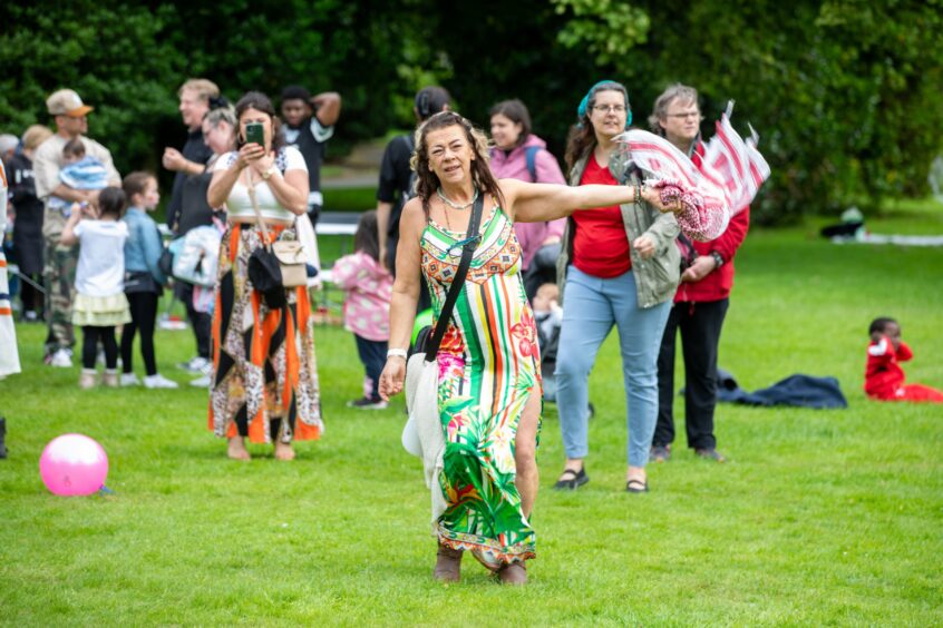 Smiling woman in a colourful dress waves a scarf while dancing on the grass among crowds at the Afro-Caribbean Summer Festival in Dunfermline.