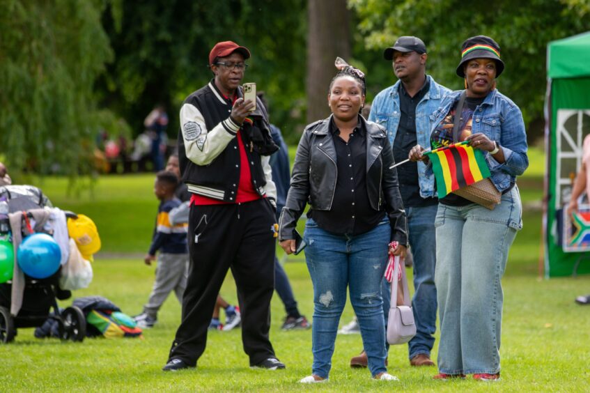 A group of attendees smiling and engaging at the Afro-Caribbean Summer Festival in Pittencrieff Park, Dunfermline, holding a Zimbabwean flag and mobile phone.
