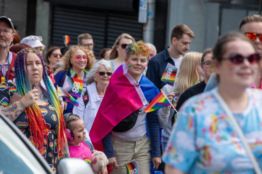 Crowd at Fife Pride 2025 in Kirkcaldy with rainbow outfits, glitter, face paint and LGBTQ+ flags, including a young person wearing a pink and blue pride cape.