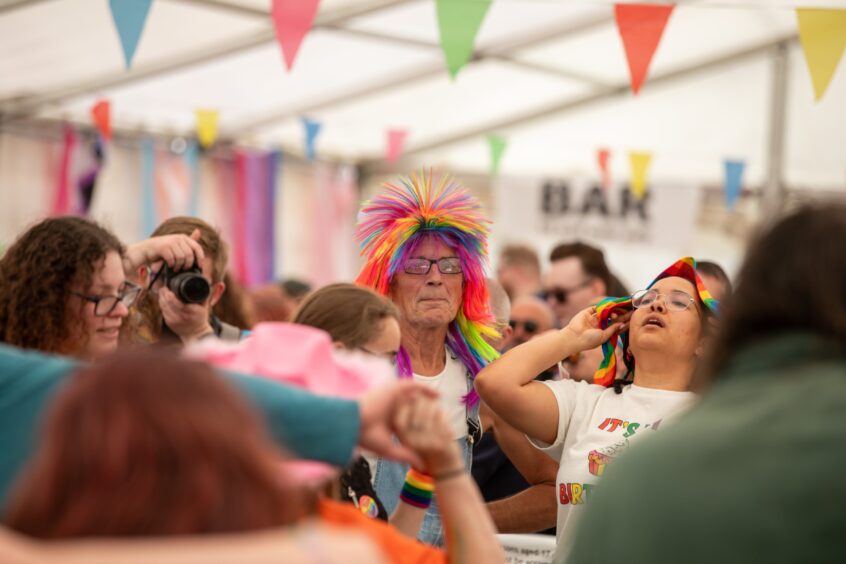 Person wearing a bright rainbow wig stands among revellers inside the Fife Pride 2025 marquee, surrounded by colourful bunting and partygoers.