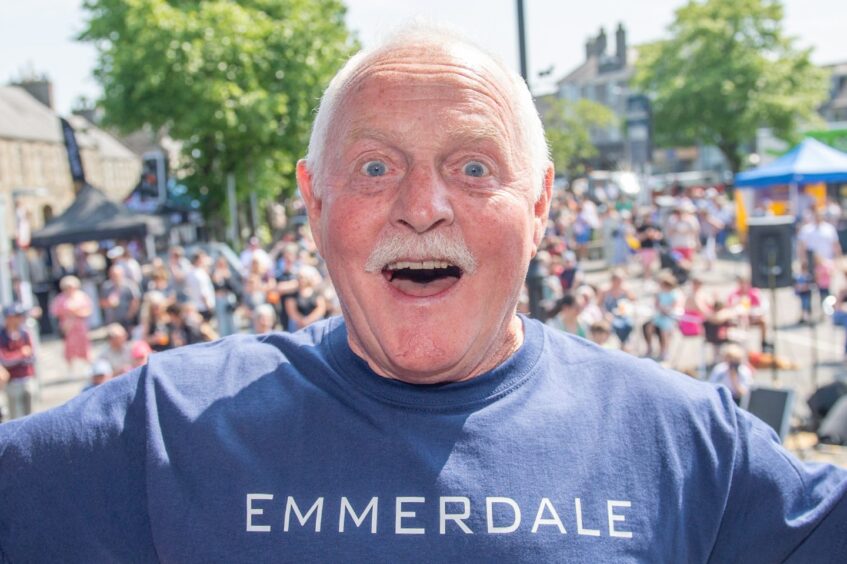 Chris Chittell grinning in Emmerdale t shirt with large outdoor crowd behind him on sunny day