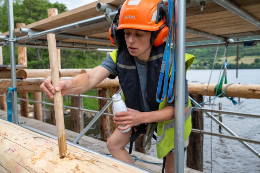 Woman in hard hat and high-vis vest inserting wooden pole into hole at crannog construction site