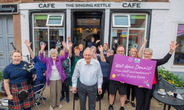 Dennis Arnold celebrates completing a walking challenge with supporters including piper Lacey Colville outside The Singing Kettle Cafe in Alyth.