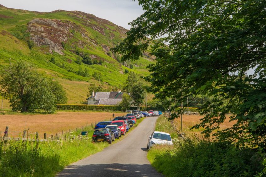Narrow track with hillwalkers' cars parked along verge and mountains in background