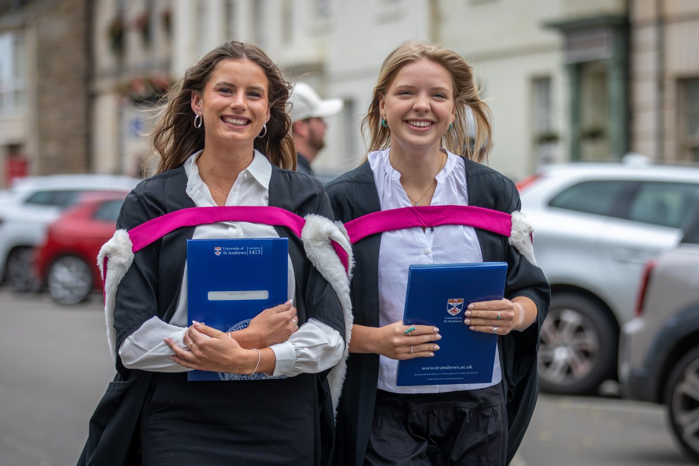 Two female graduates in black gowns with pink hoods walk side by side, smiling and holding blue University of St Andrews folders, with parked cars in the background.
