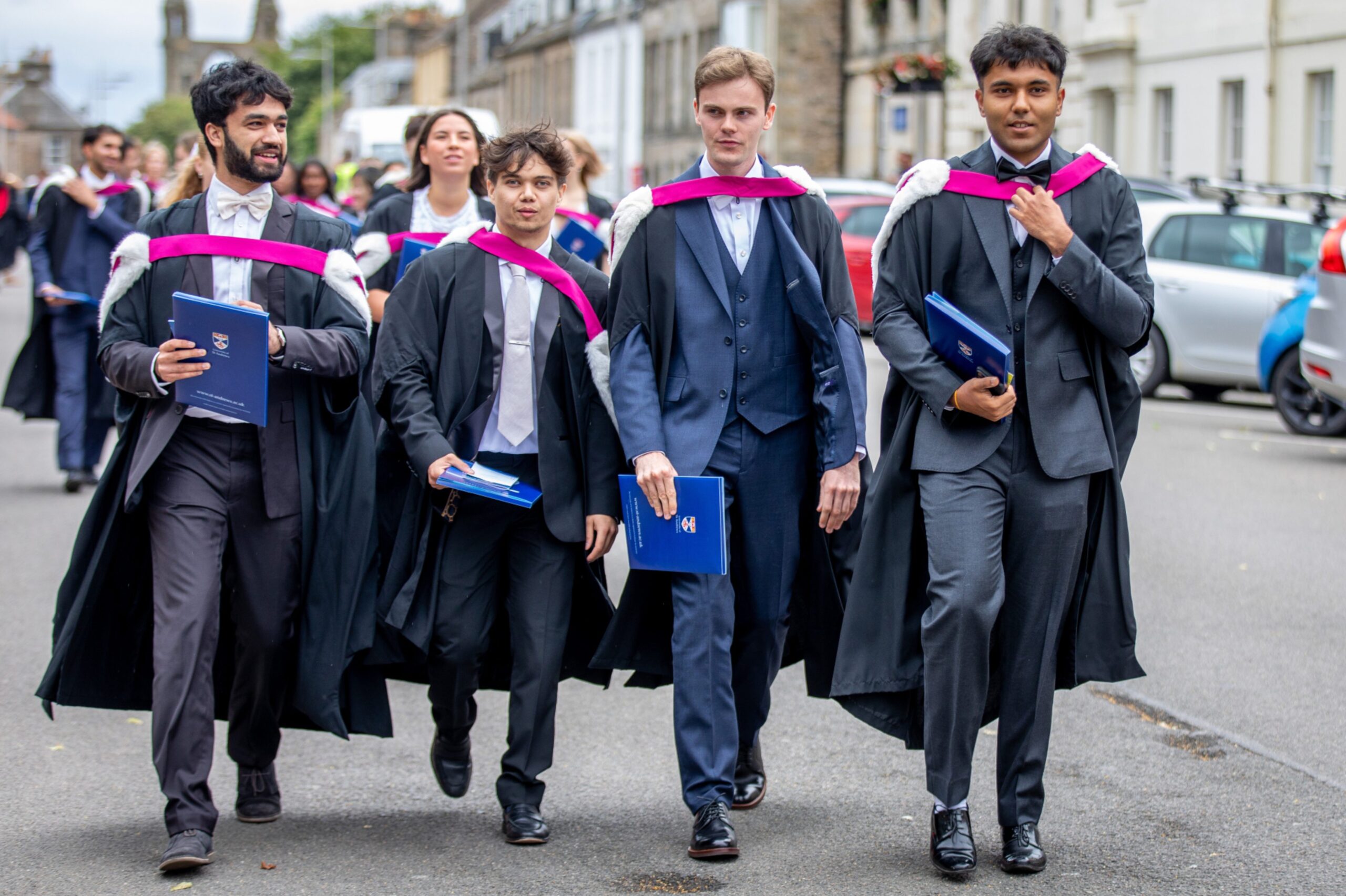 Four male graduates in dark suits and academic gowns walk down a street, holding blue St Andrews degree folders, with cars and buildings lining the background.