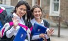 Two female graduates in gowns and white shirts smile and wave while holding blue University of St Andrews folders during the graduation parade.