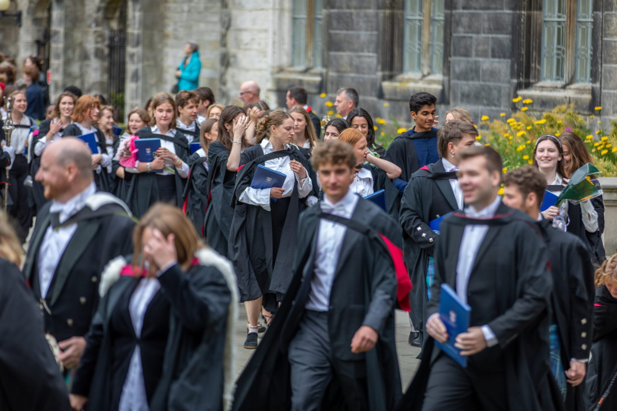 Large group of graduates in gowns and hoods walk through the university grounds at St Andrews, many smiling and chatting as they head toward St Salvator’s Quad.