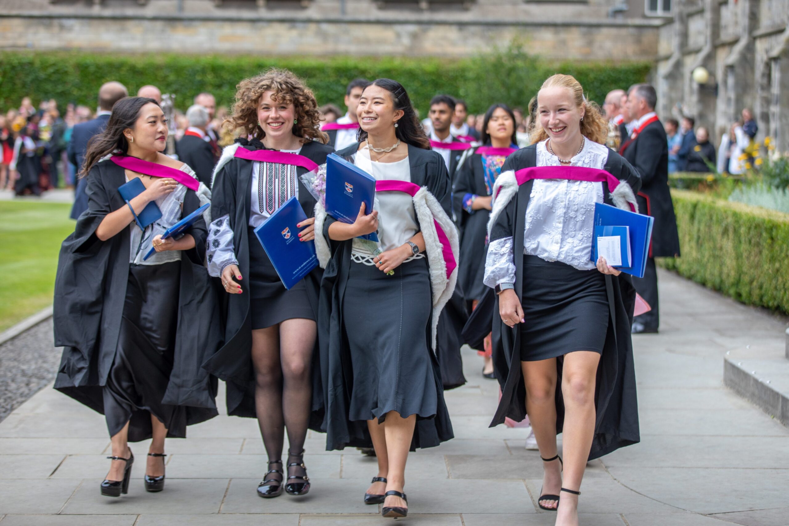 Four smiling graduates walk arm-in-arm in black gowns with pink and white hoods, holding diplomas as they leave Younger Hall for St Salvator’s Quad at the University of St Andrews.