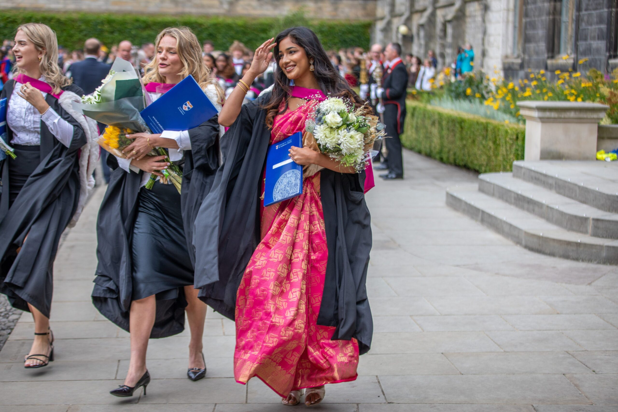 Three graduates in academic gowns, including one in a pink sari, walk with flowers and degree folders during the St Andrews University graduation parade.
