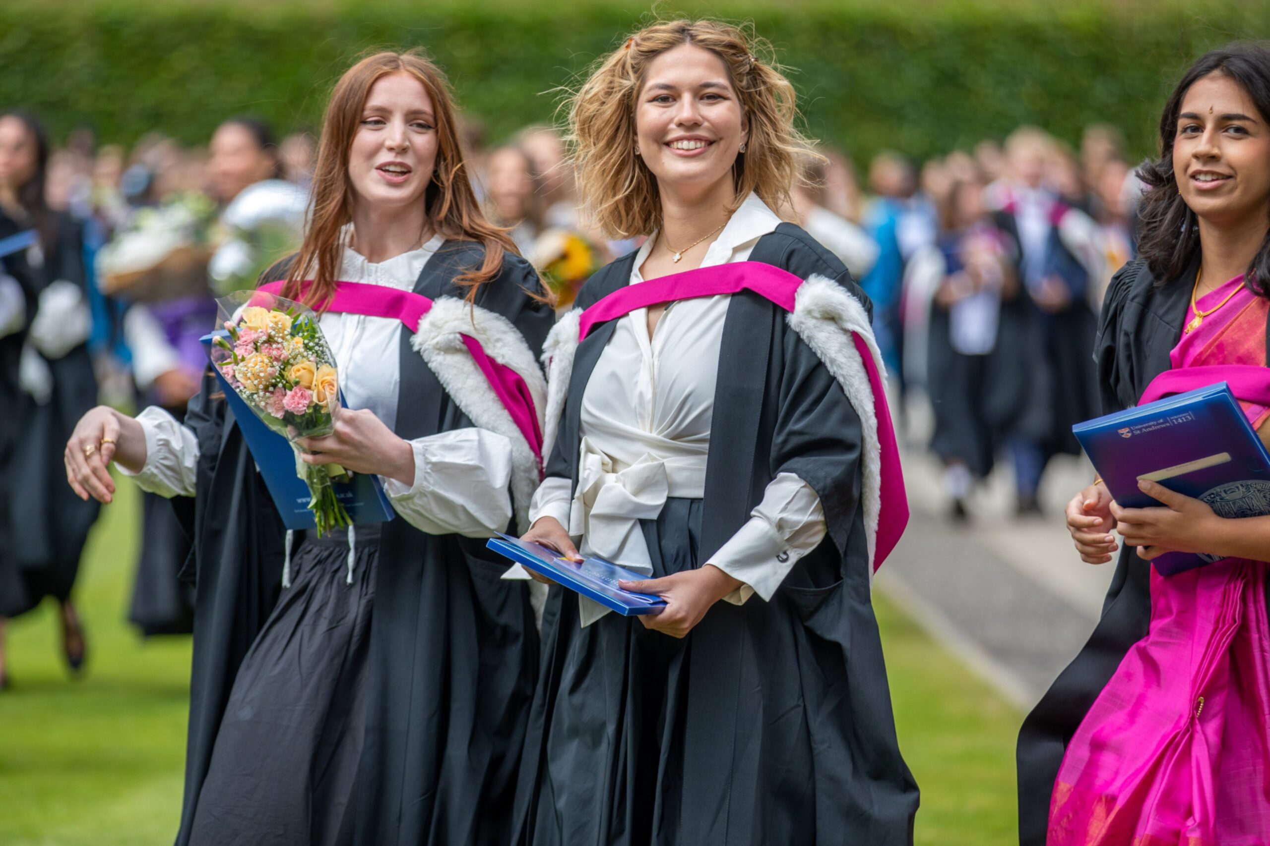Three female graduates in gowns, one in a pink sari, walking together with flowers and certificates between Younger Hall and St Salvator’s Quad.