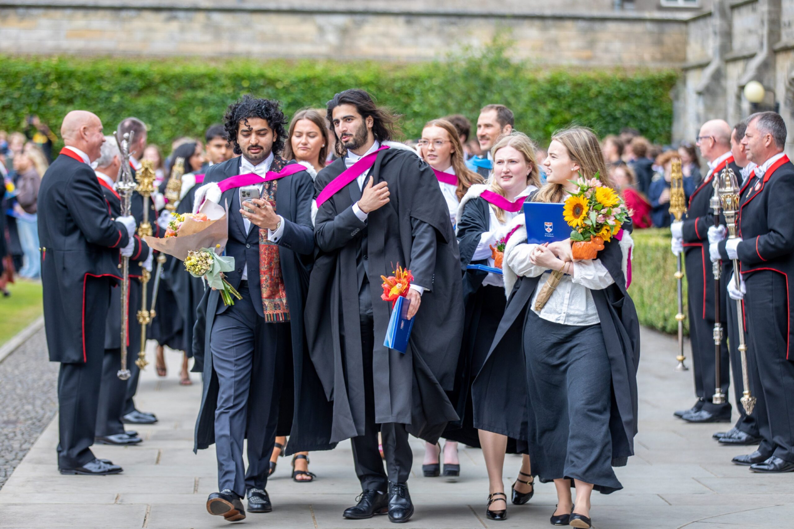 Graduates in gowns walking from Younger Hall to St Salvator’s Quad, some holding flowers and phones, flanked by ceremonial staff.
