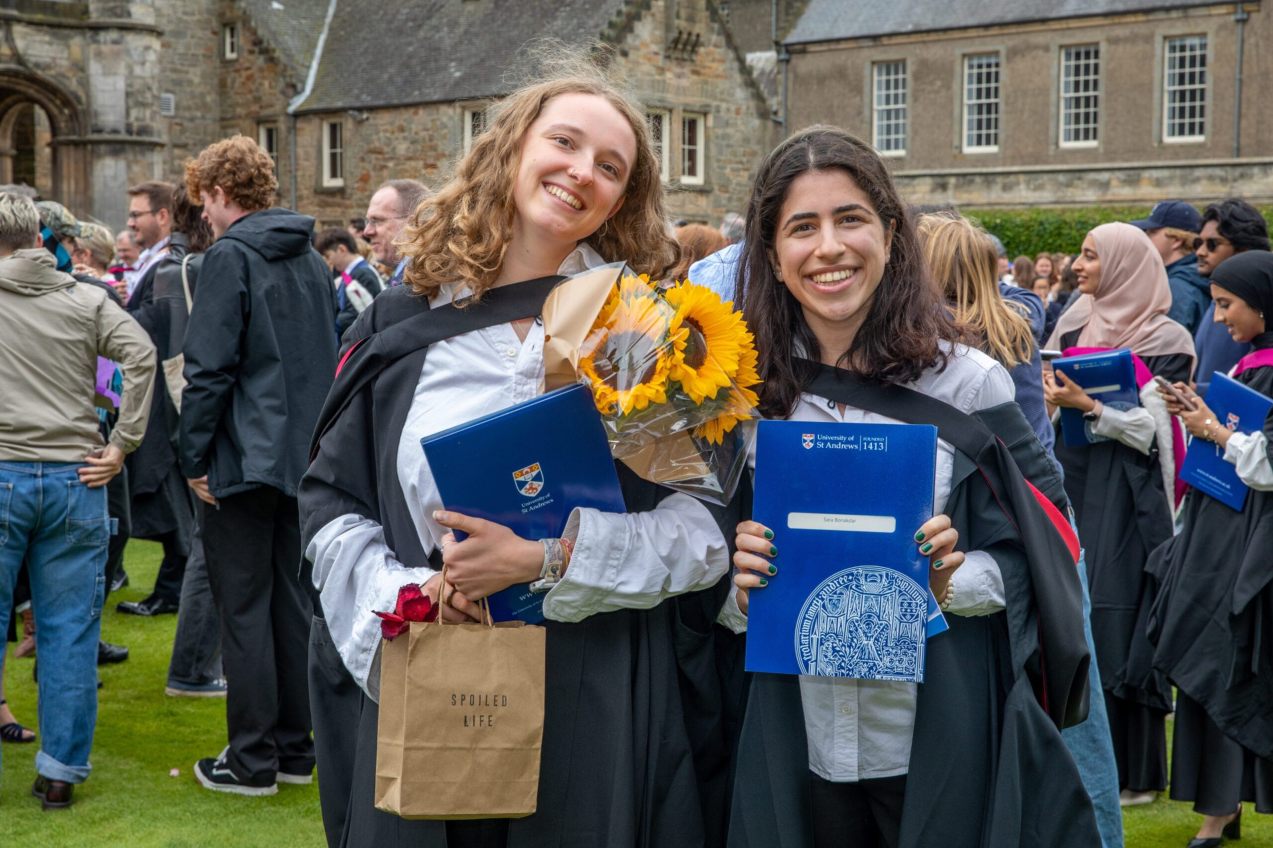 Two smiling graduates with gowns and folders stand together in St Salvator’s Quad, one holding sunflowers and a gift bag.