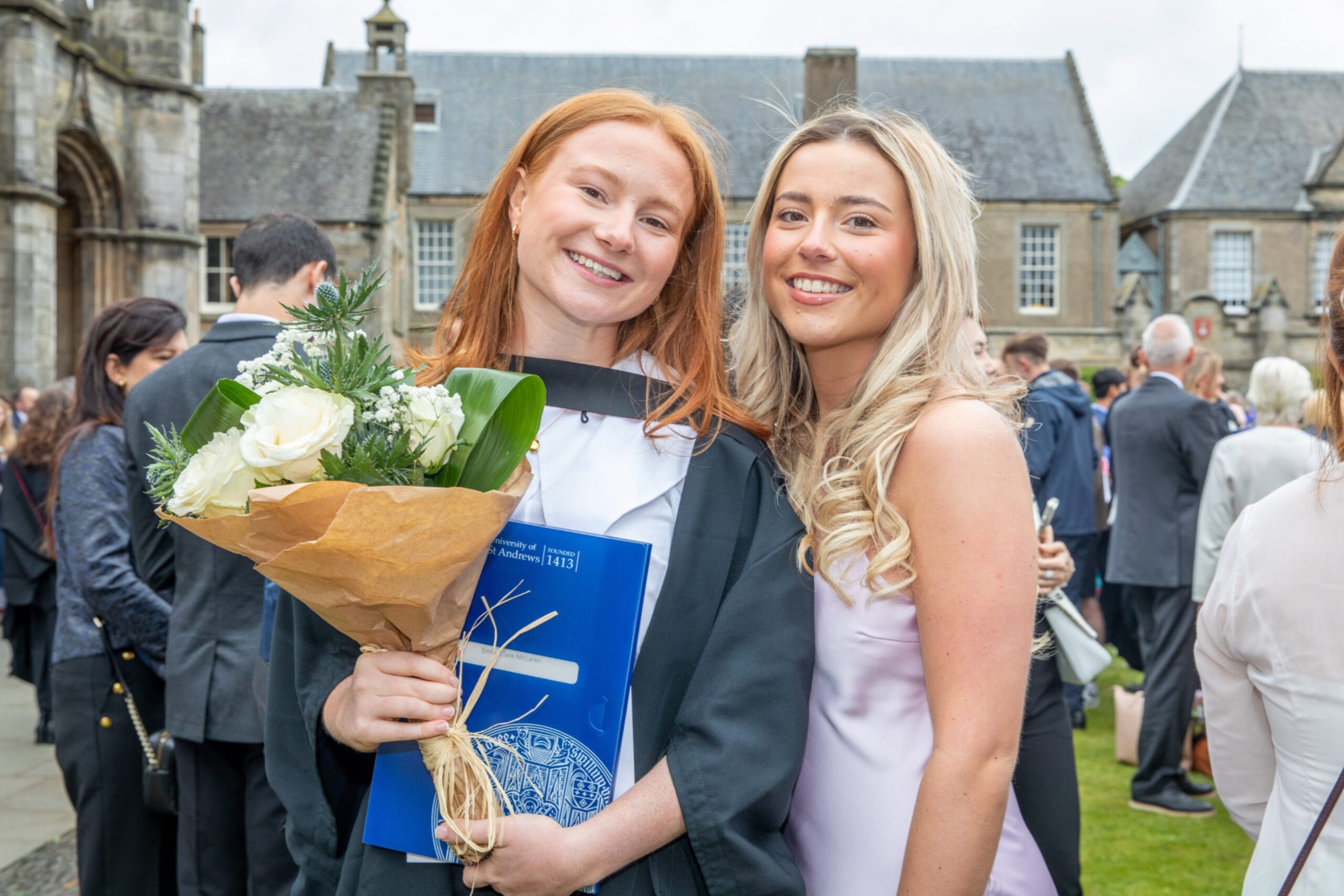 Graduate Emily McLaren with sister Sophie McLaren, both smiling and holding flowers in St Salvator’s Quad at the University of St Andrews.