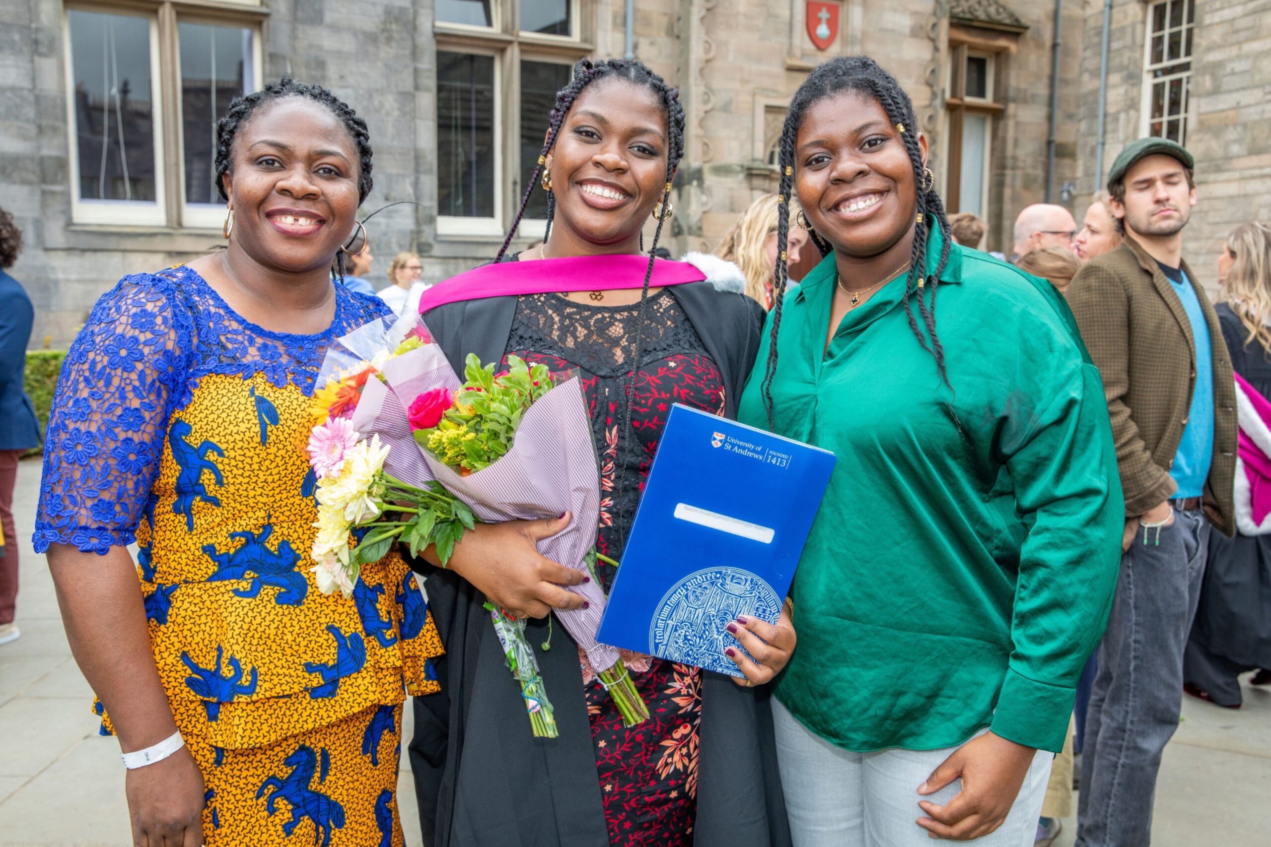 Graduate Naomi Omorogbe Alakiri holding flowers and a certificate, smiling with Ebunollwa Alarkiri Edokpayi and Sarah Omorogbe Alakiri in St Salvator’s Quad.