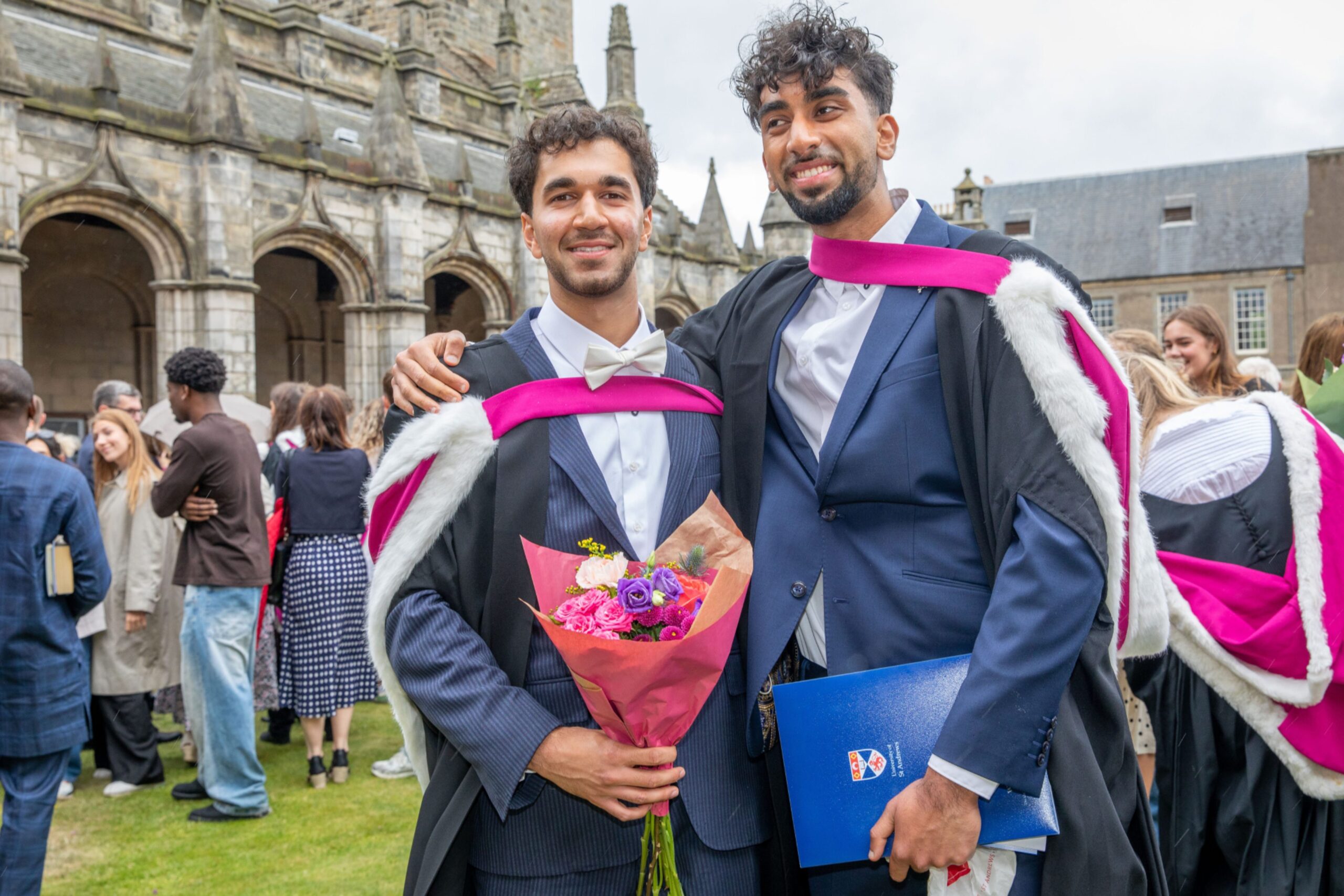 Graduates Musa Tariq and Ken Kont in gowns, smiling with flowers and certificate folders in St Salvator’s Quad after graduating from St Andrews.