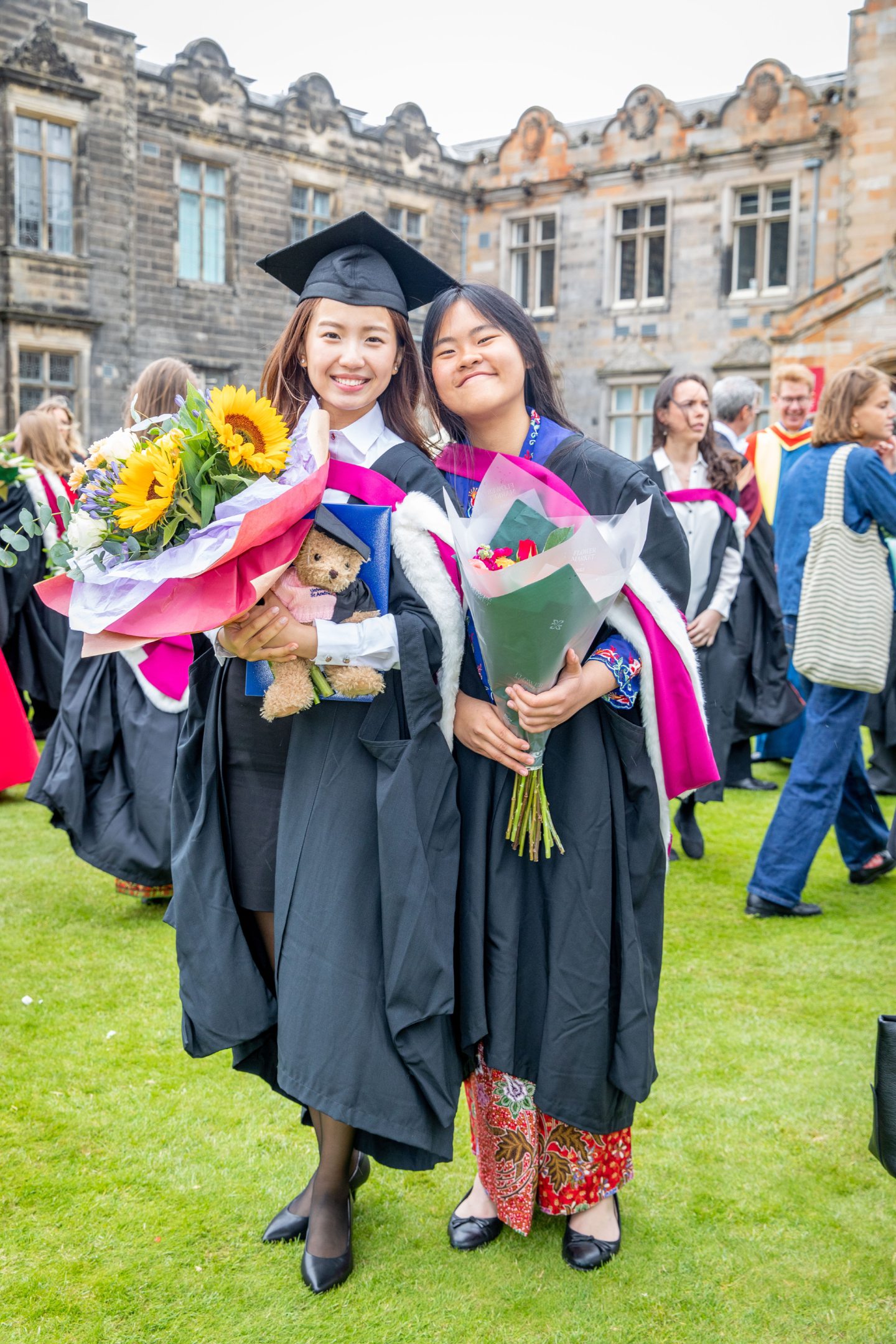 Graduates Wing Yi Chilli Lam and Lim Pei Yi smiling in gowns with flower bouquets and folders in St Salvator’s Quad, St Andrews.