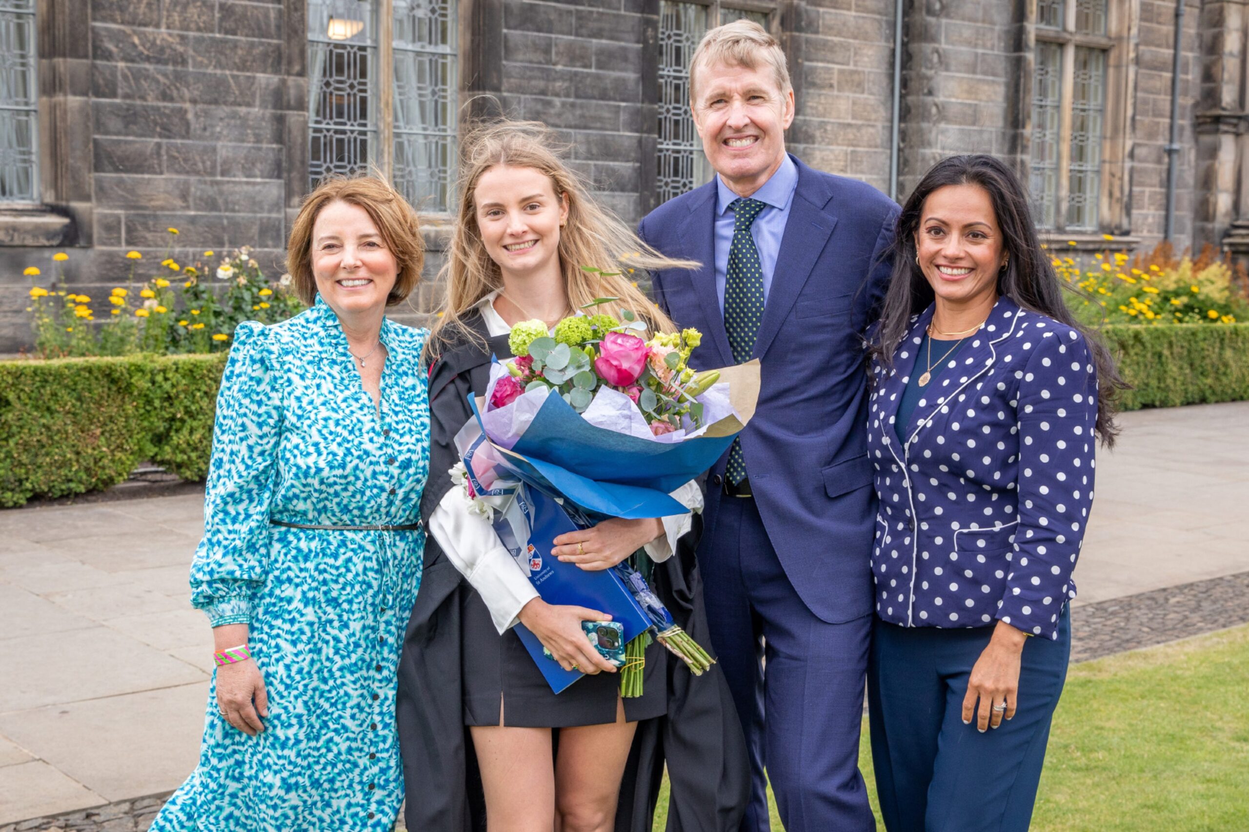 Graduate Amy Bartholemew holding flowers and standing with her smiling family in front of a historic stone building at St Andrews.