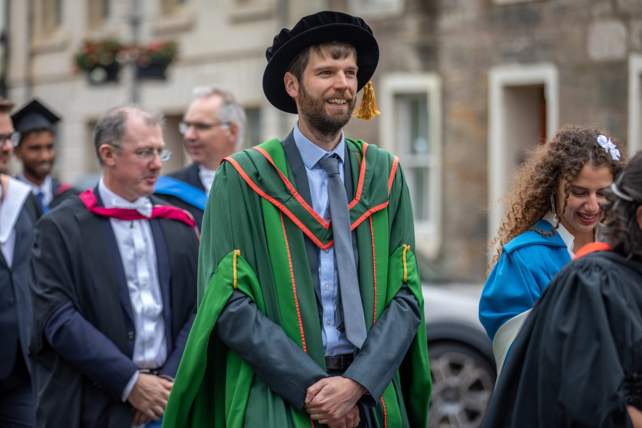 Smiling academic in green and orange robes leads a procession from Younger Hall to St Salvator’s Quad during the St Andrews graduation parade.