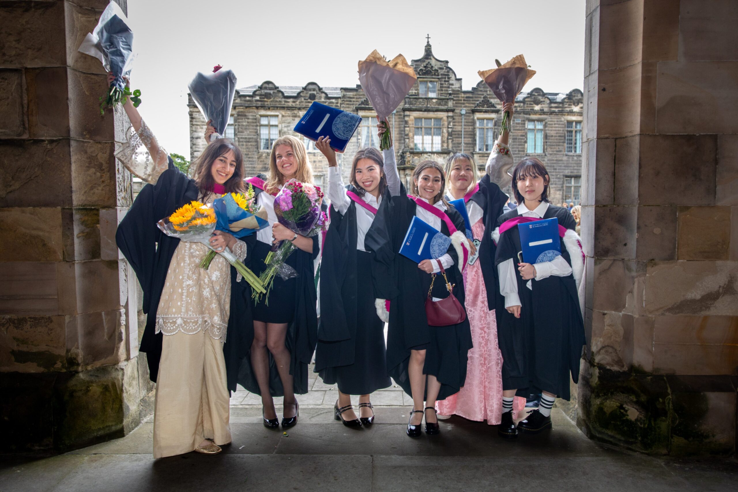 Graduates Haniyah Khan, Aurelia Tiffoney, Sarah Ng, Liana Motamedi, Jimin Lim and Hyomin Jung posing with flowers and degree folders under a stone arch in St Andrews.