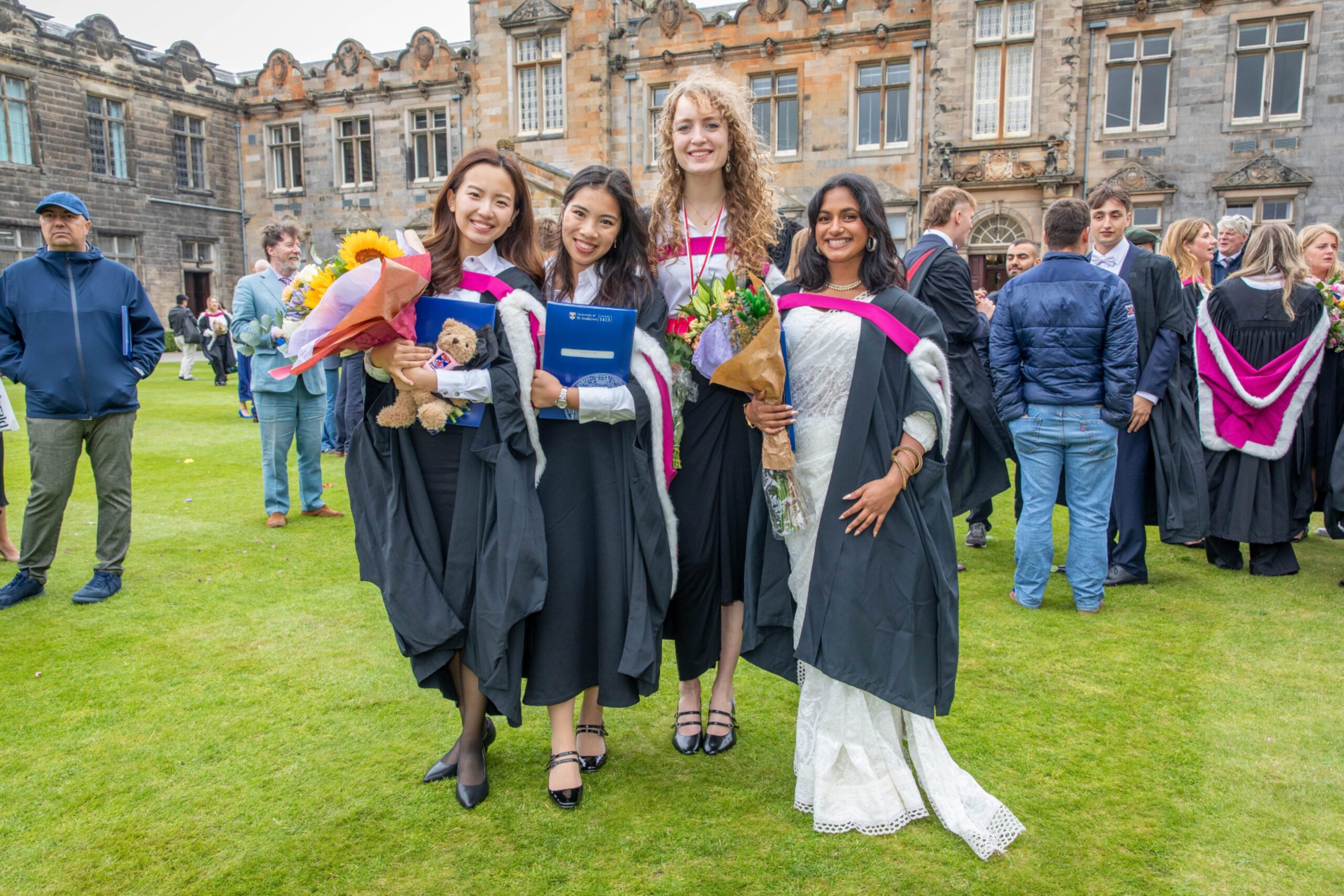 Graduates Chilli Lau, Sarah Ng, Lola Chirico and Chriswina Lobo smiling and holding bouquets in front of St Andrews’ historic buildings.