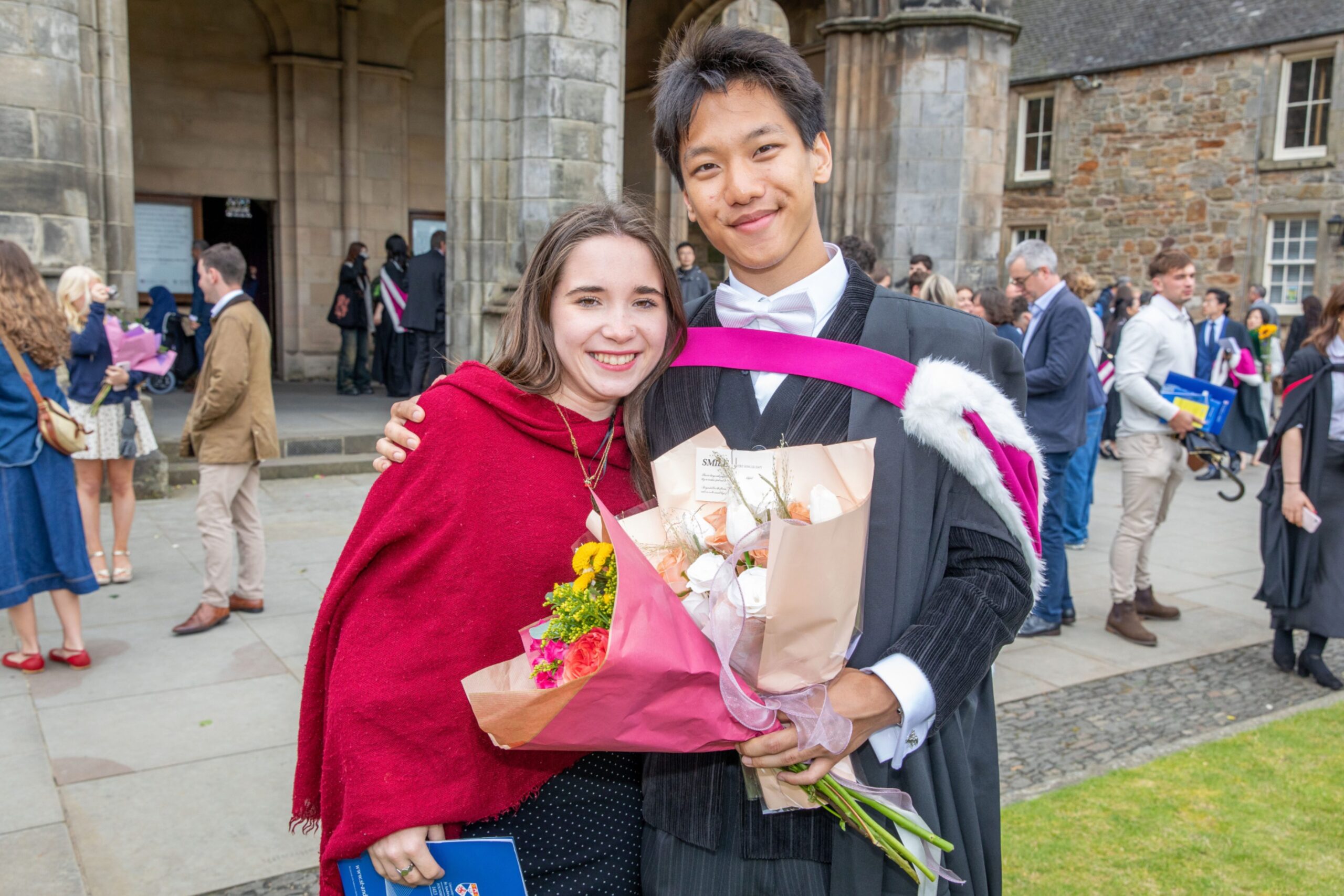 Graduate Godwin Choi with Verity Wilson, both smiling and holding bouquets outside Younger Hall after the Friday morning ceremony in St Andrews.