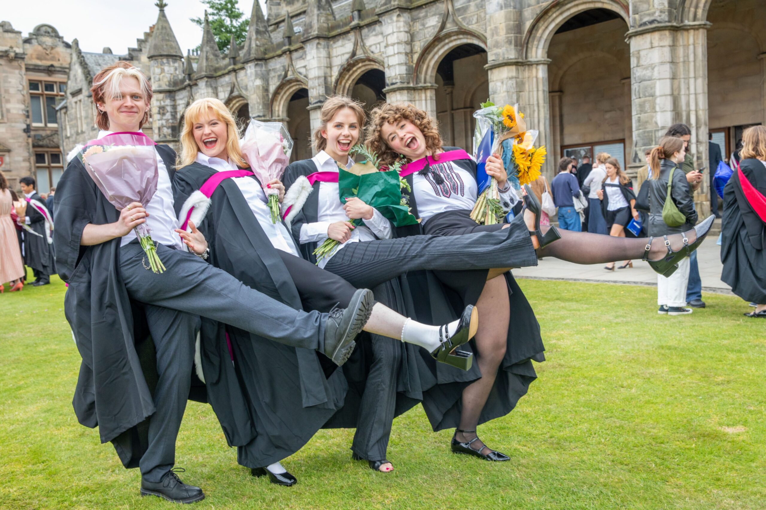 Graduates Tom Hilton, Charlie Williamson, Karen Jakobsdottir and Rosalia Kancir pose with high kicks and flowers outside Younger Hall in St Andrews.