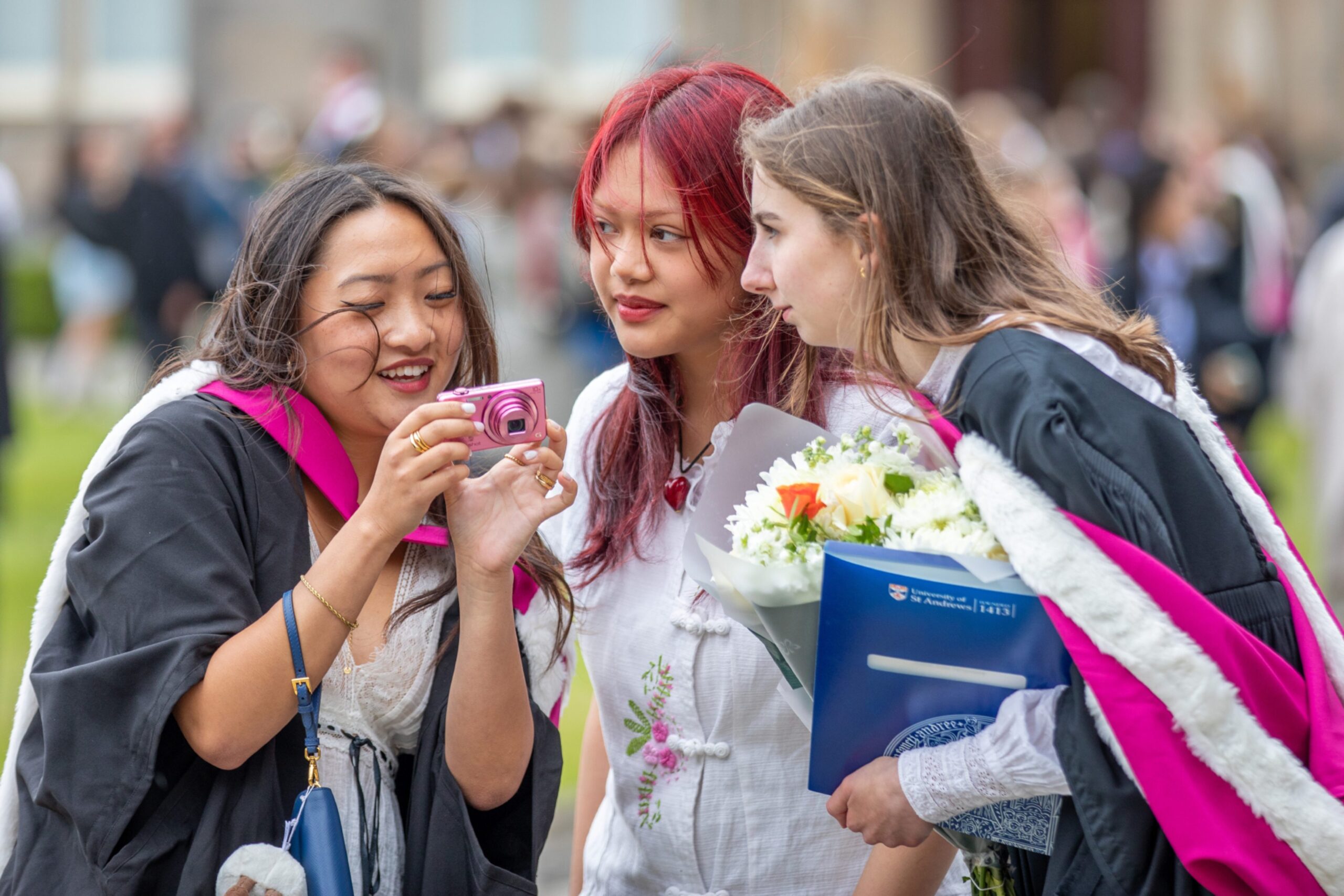 Three young women, including two graduates in gowns, looking at a pink camera and smiling during St Andrews graduation day.