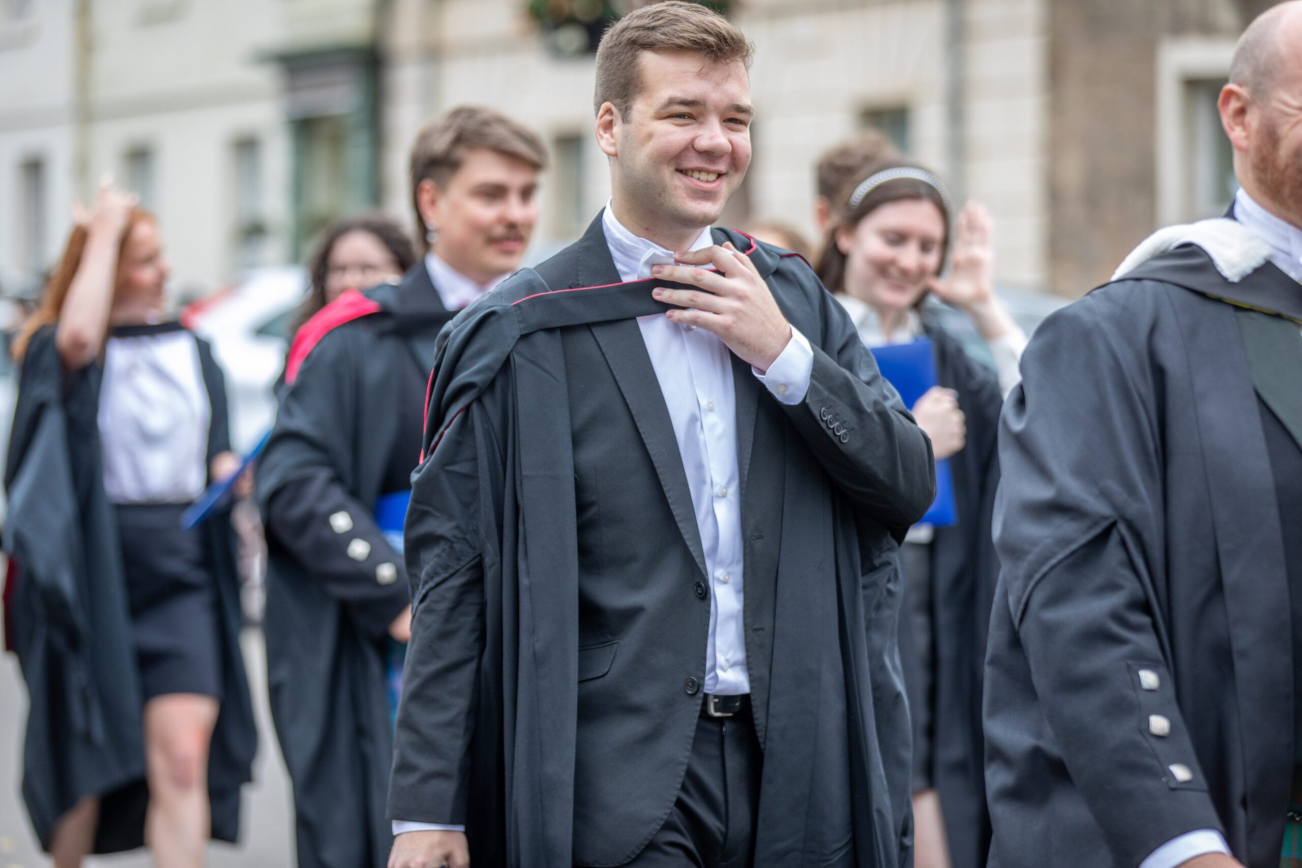 Male graduate smiling in gown while walking along South Street, St Andrews, after the Friday morning graduation ceremony.