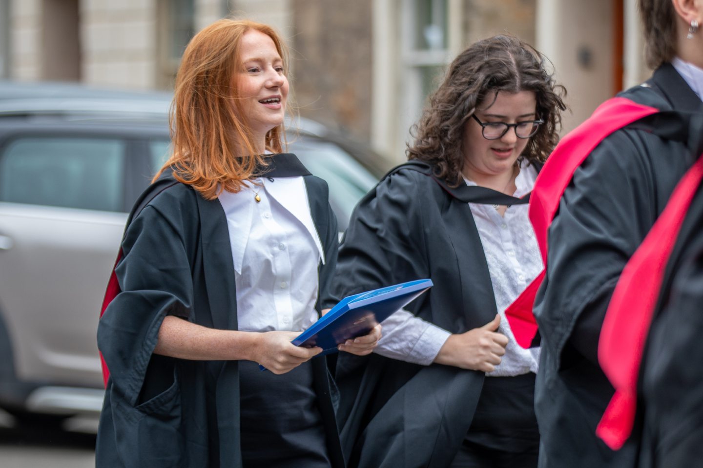 Two smiling graduates in gowns walking along South Street, St Andrews, after the Friday morning graduation ceremony.